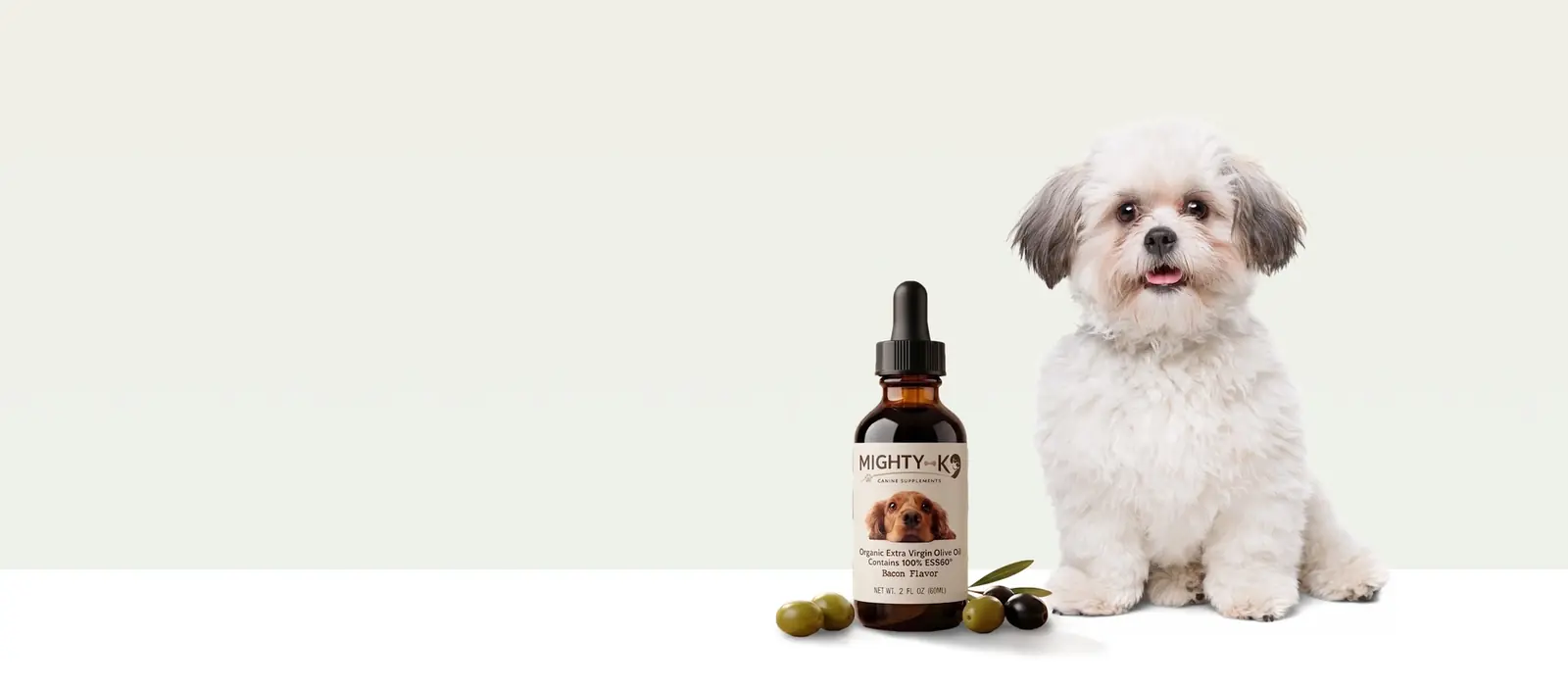 A small white fluffy dog sitting beside a Mighty K9 bottle and a few olives on a pale background
